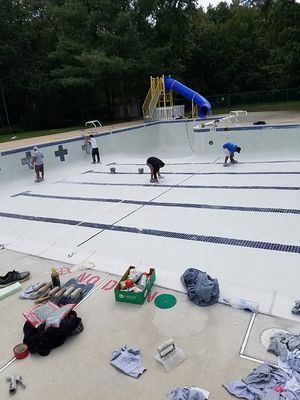 Workers repair a drained pool. They are painting, with tools and supplies scattered around. A slide is visible in the background.