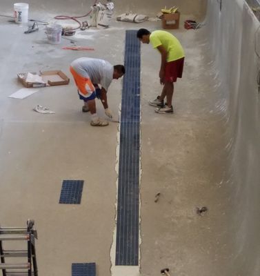 Two people installing tile in a light-colored pool under construction. One person is kneeling, applying something to the tile.