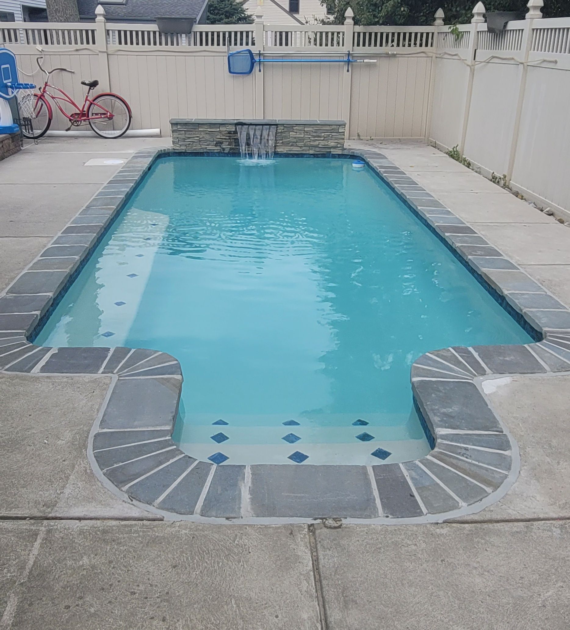 Rectangular swimming pool with waterfall feature, bordered by gray pavers. A pink bicycle rests nearby.