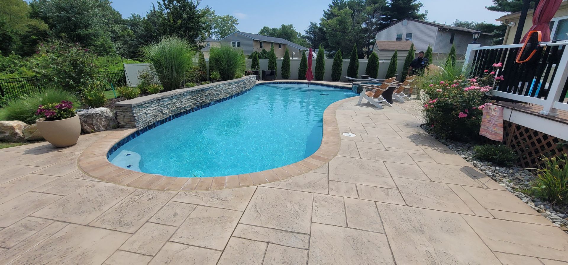 A pool surrounded by tan stone with lush greenery and a deck on a sunny day.