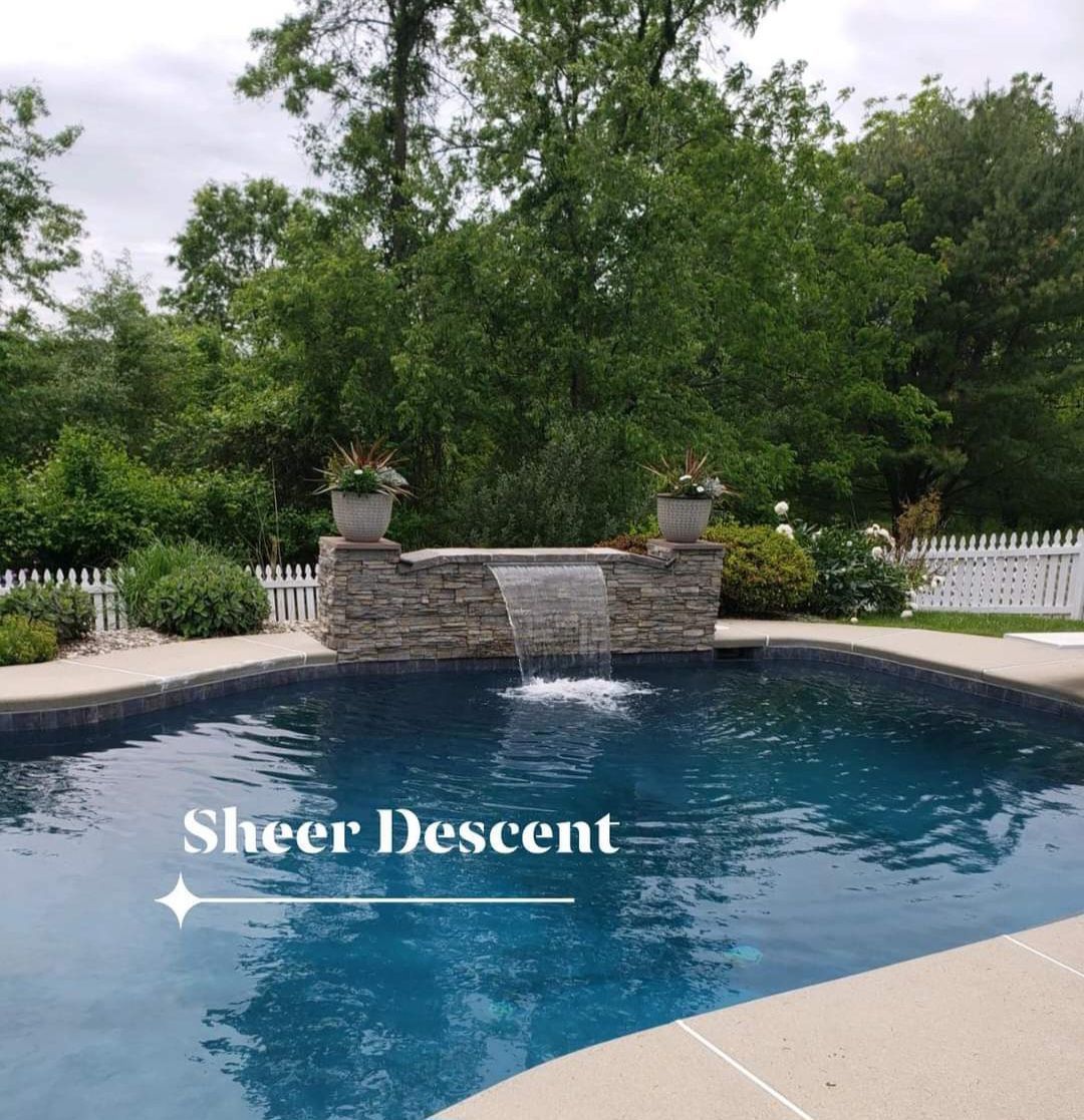Swimming pool with a stone wall water feature and two potted plants; trees in the background.