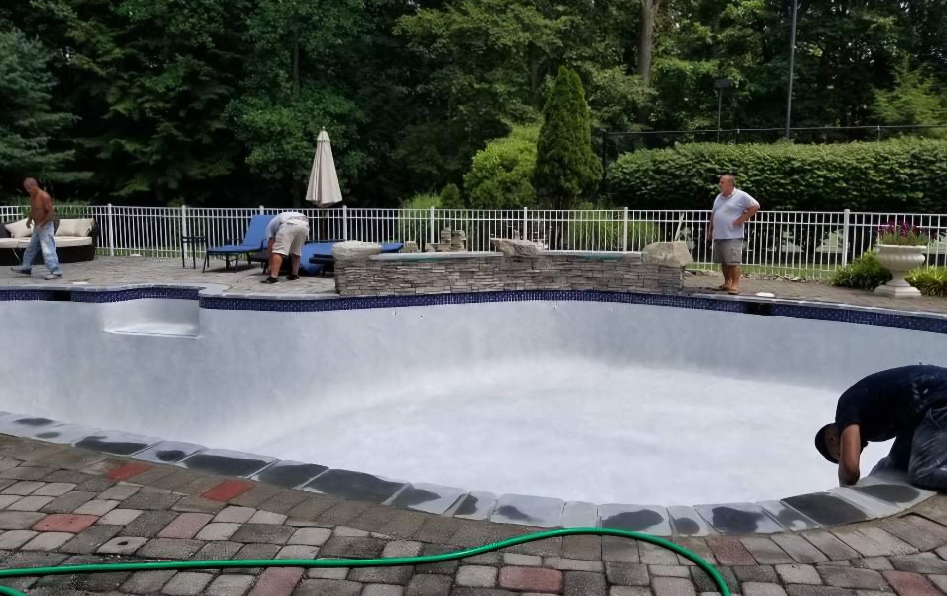Men working on an empty pool with blue tile trim. Poolside, green hose, trees, and white fence in background.