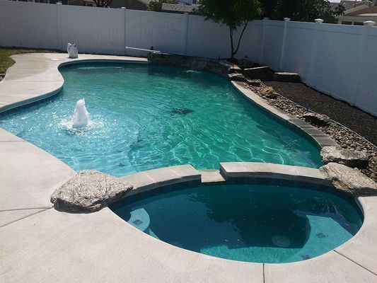 Swimming pool with fountain and connected spa, surrounded by concrete and white fence.