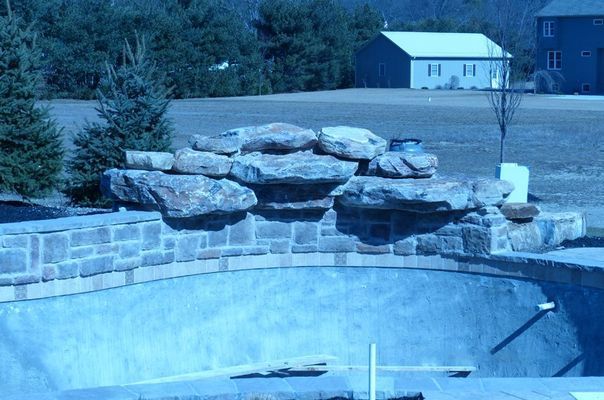 Unfinished swimming pool with stacked rock waterfall feature. Outdoors, clear sky.