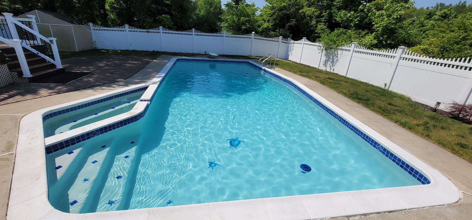 Swimming pool surrounded by a white picket fence, steps leading into the water, and lush greenery.