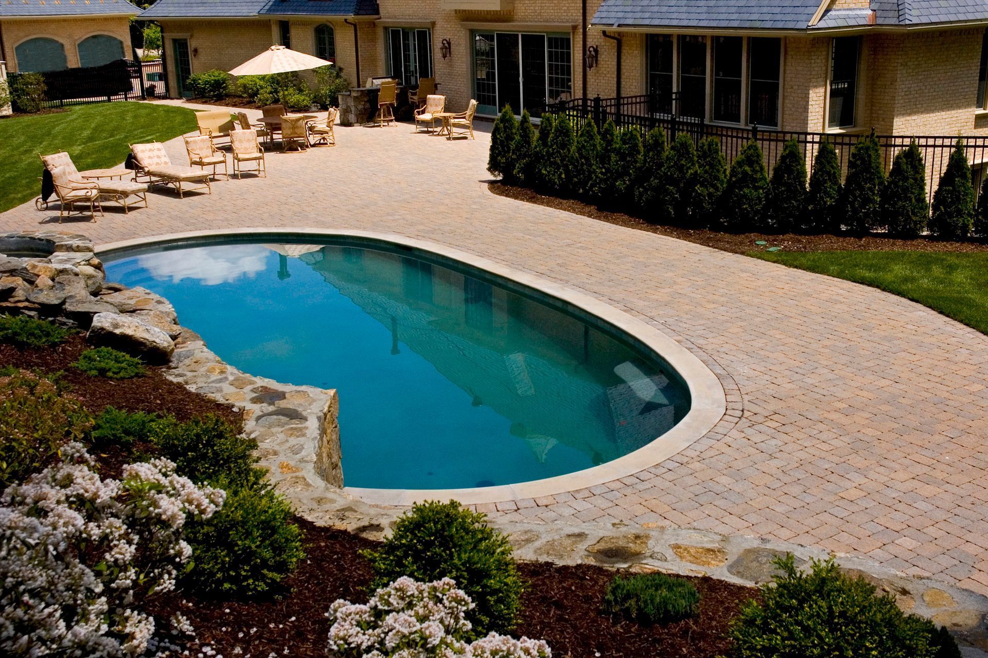 Swimming pool with stone edging and brick patio, surrounded by landscaping and a house.