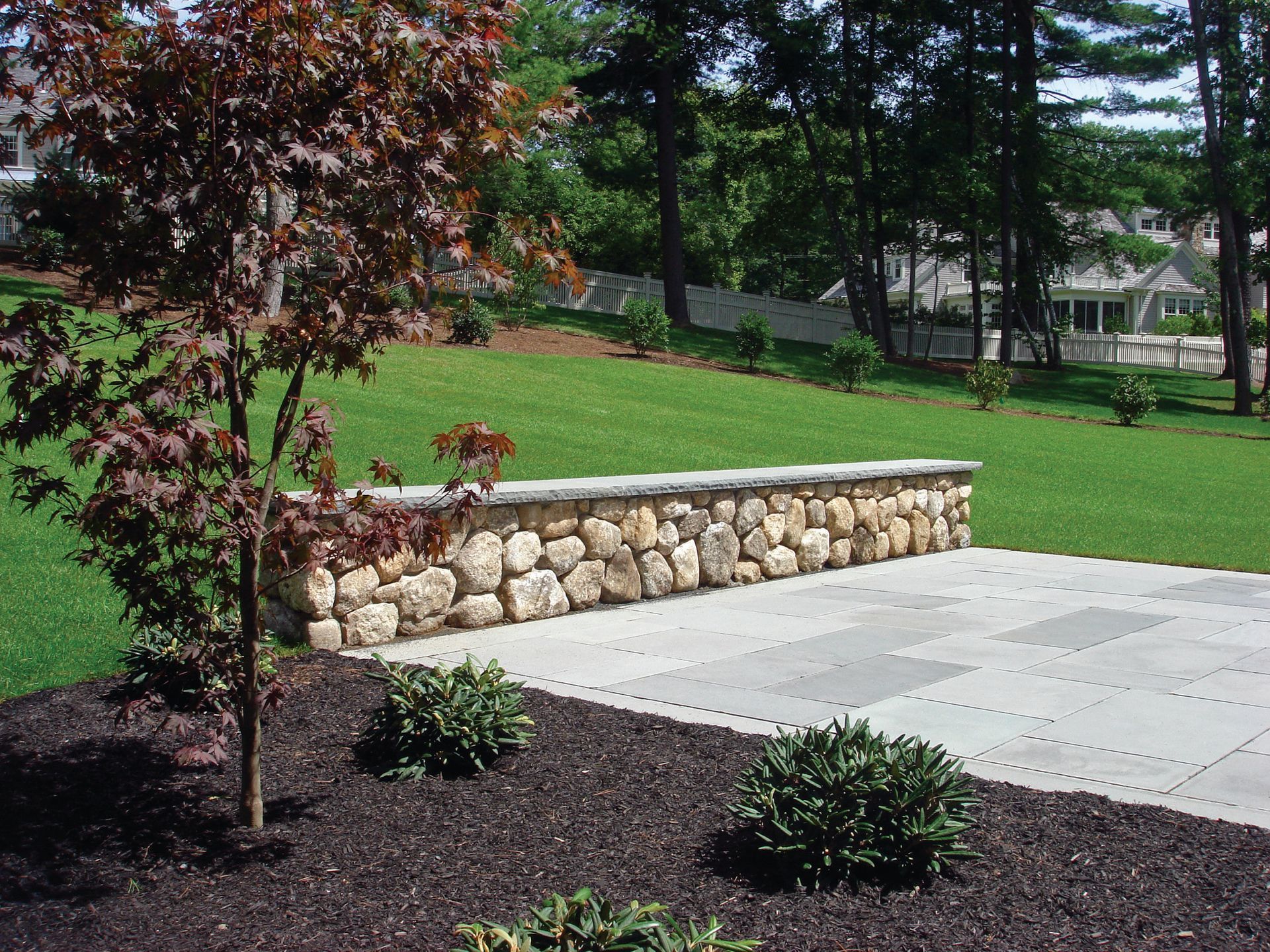Patio with stone wall and landscaping; tree on left, green lawn in background.