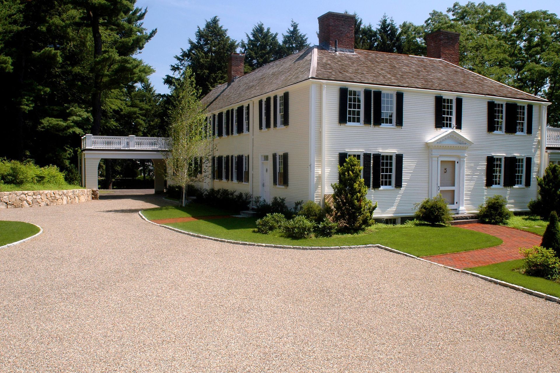 Two-story white colonial house with black shutters and a gravel driveway, trees in background.