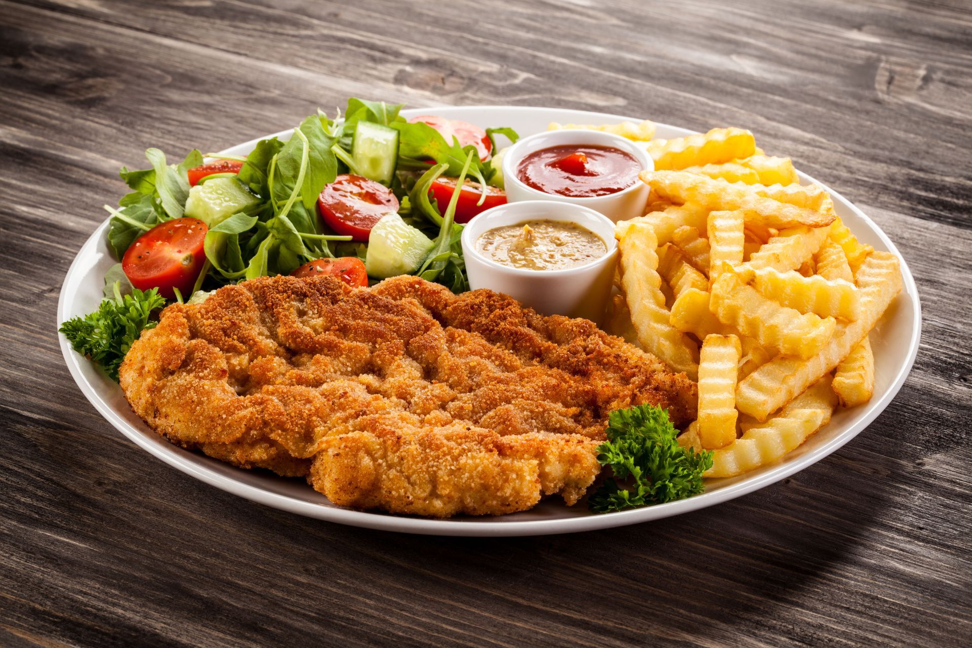 Plate of fried cutlet, fries, salad, and dipping sauces on a wooden table.