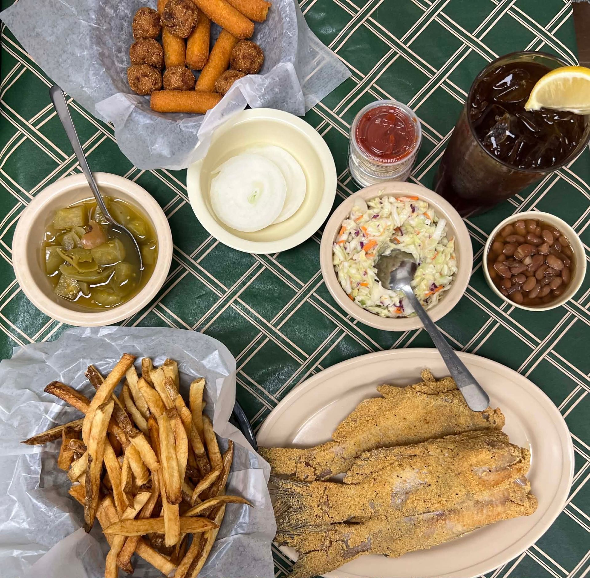 Overhead view of a meal: fried fish, fries, beans, slaw, onion rings, hushpuppies, tea, and sauces on a checkered tablecloth.
