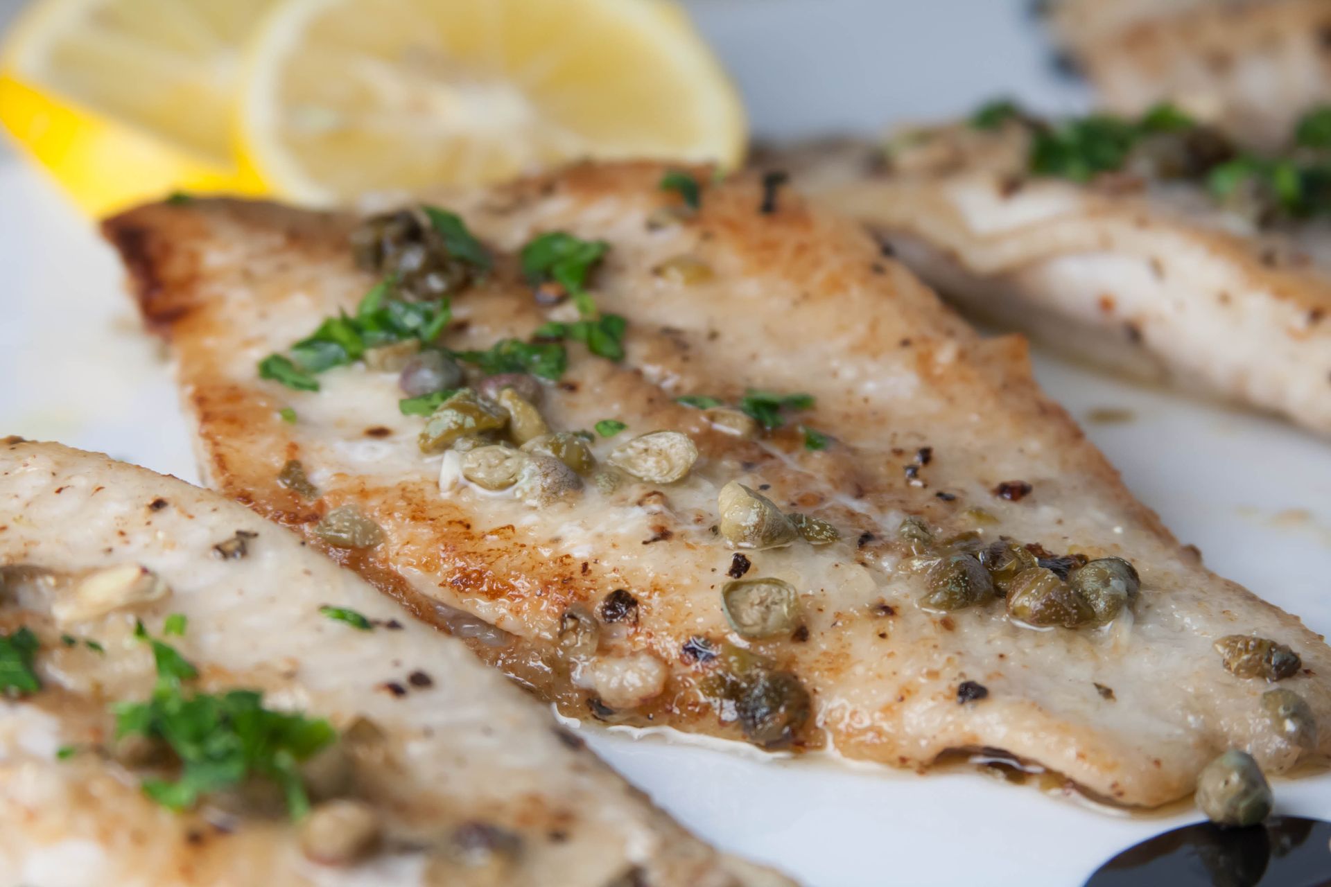 Pan-seared fish fillets with capers, parsley, and lemon wedges on a white plate.
