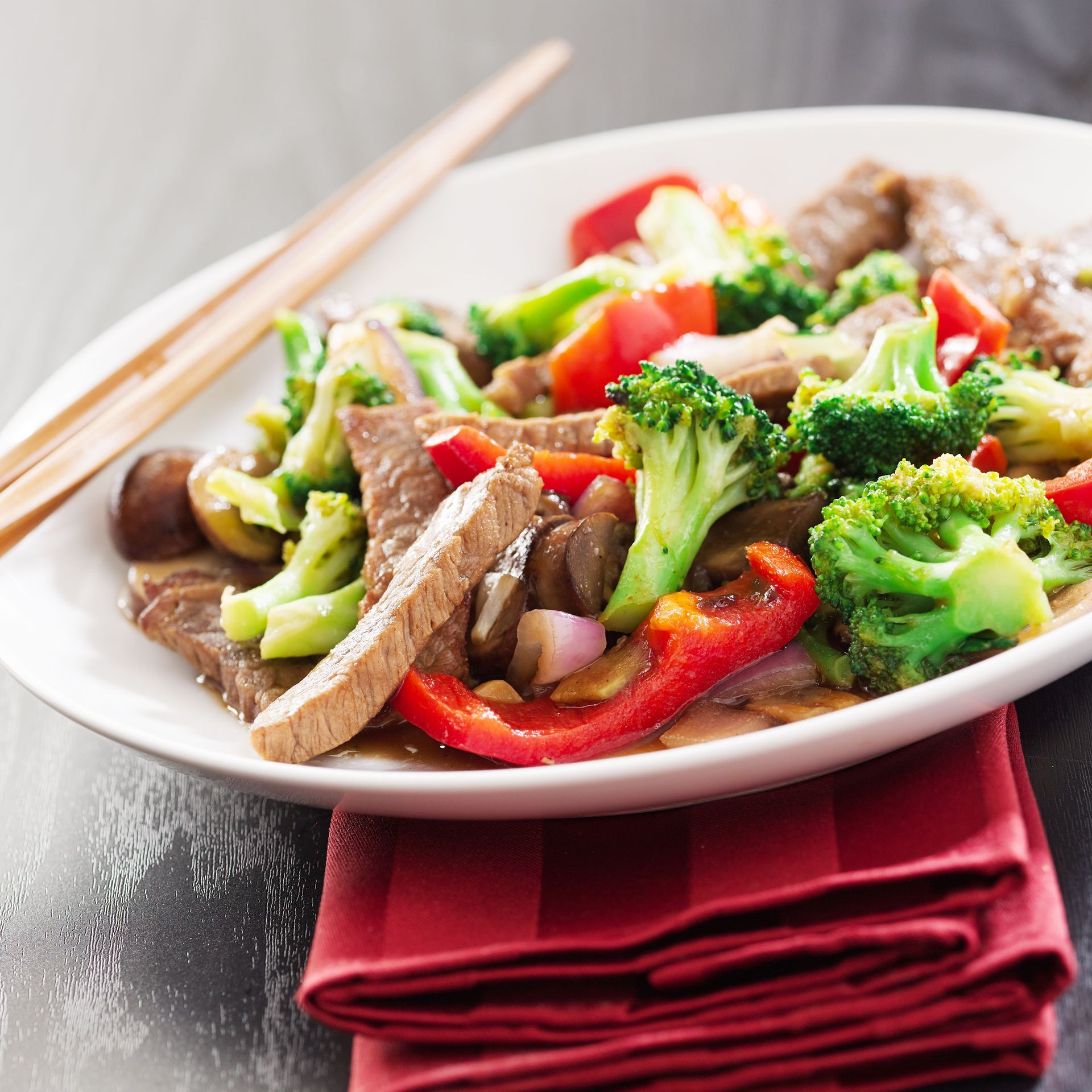 Beef and broccoli stir-fry with red peppers, served on a white plate with chopsticks and red napkin.