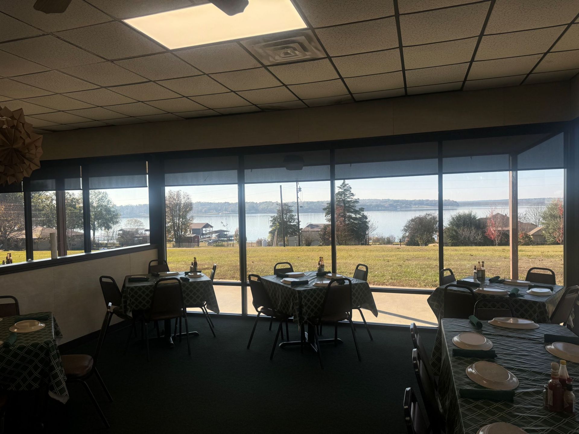 Restaurant interior with tables set for dining, large windows overlooking a lake and green lawn.