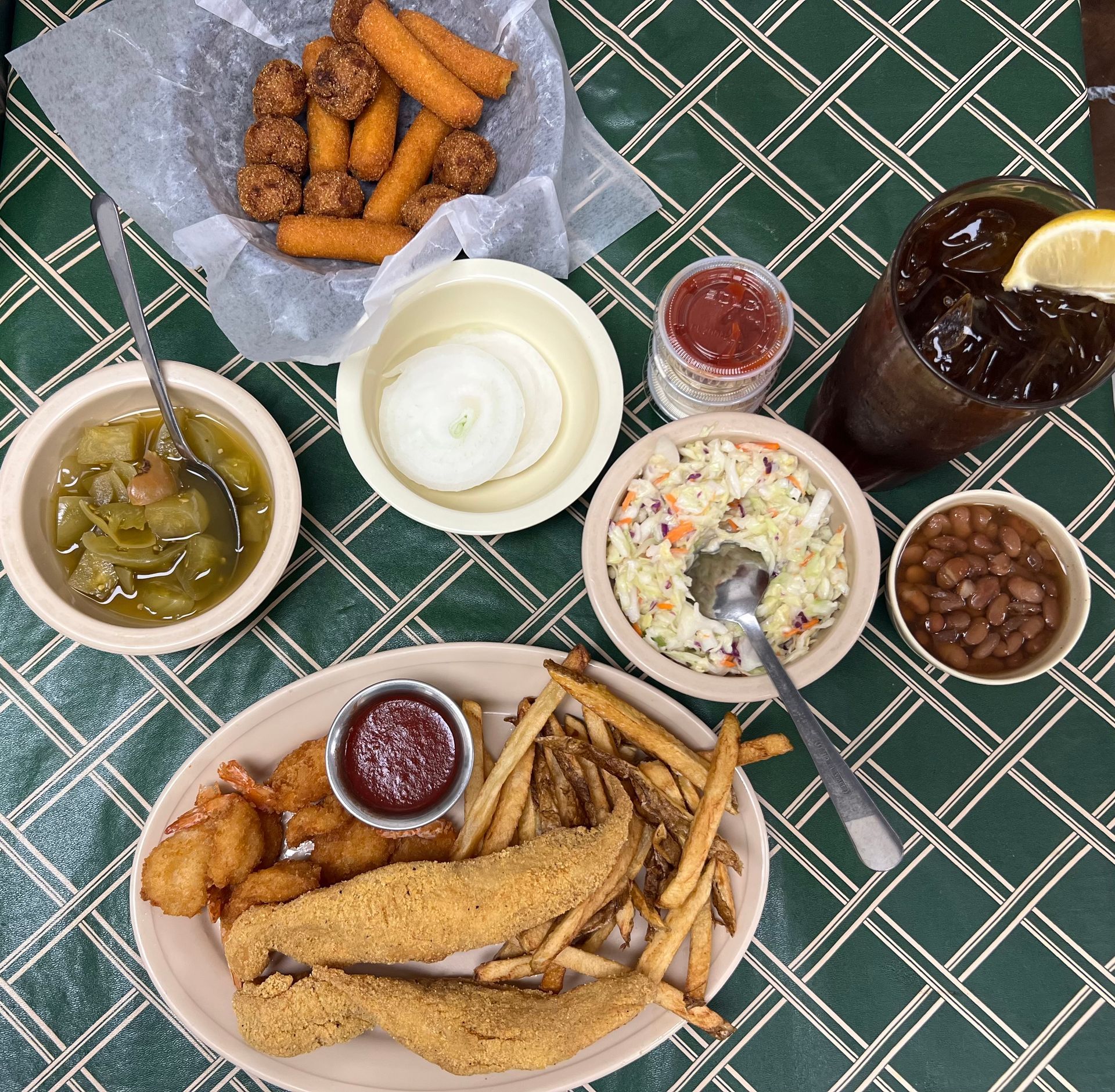 A table laden with Southern comfort food: fried fish, fries, coleslaw, beans, onion, and a drink.