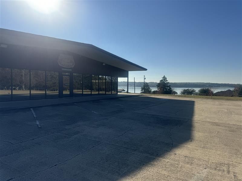 A single-story building with large windows and a slanted roof sits in front of a body of water under a bright sun.