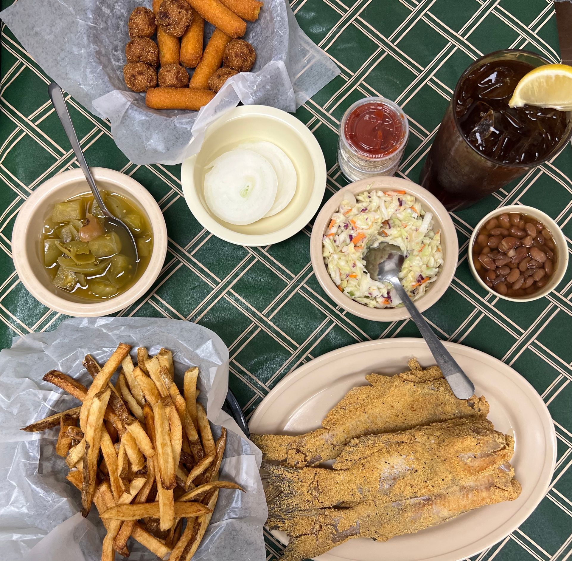 Fried fish, fries, and sides on a checkered tablecloth, including coleslaw, beans, onion rings, and a drink with lemon.