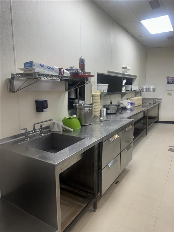 Stainless steel commercial kitchen with a sink, prep surfaces, shelves, and storage.