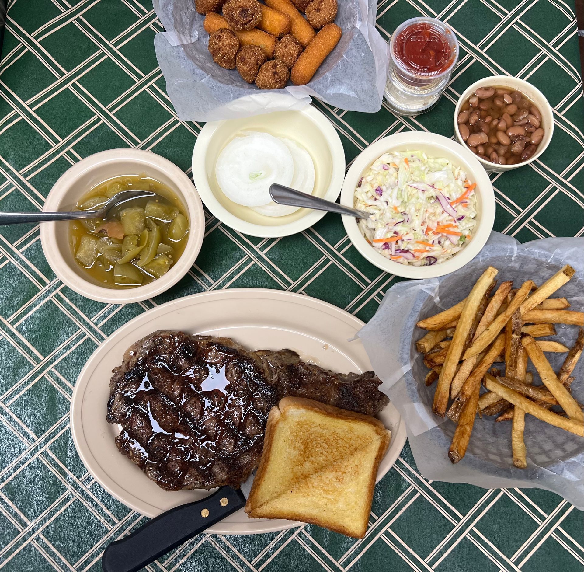Steak dinner with sides: fries, coleslaw, beans, vegetables, corn fritters, toast, and sauce on a green-checkered tablecloth.