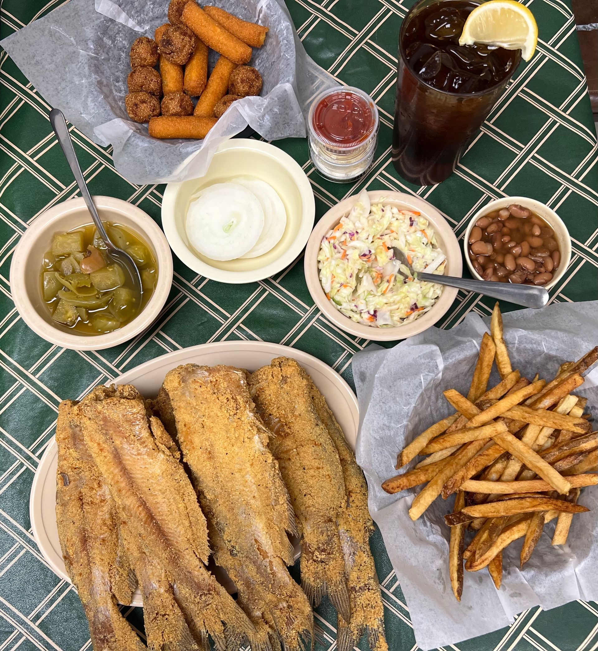 A plate of fried fish with sides of fries, coleslaw, beans, and drinks on a patterned table.