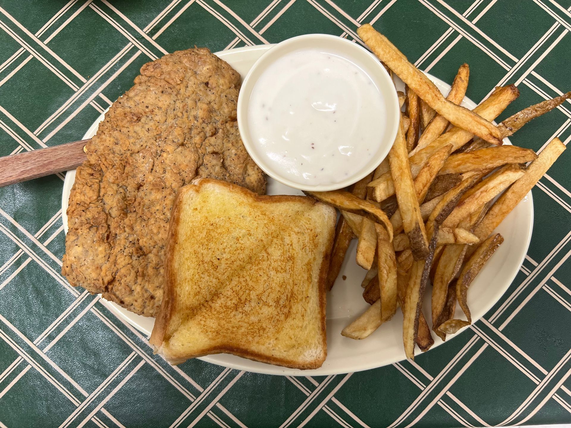 Plate with breaded steak, toast, fries, and a bowl of creamy sauce on a green-checkered surface.