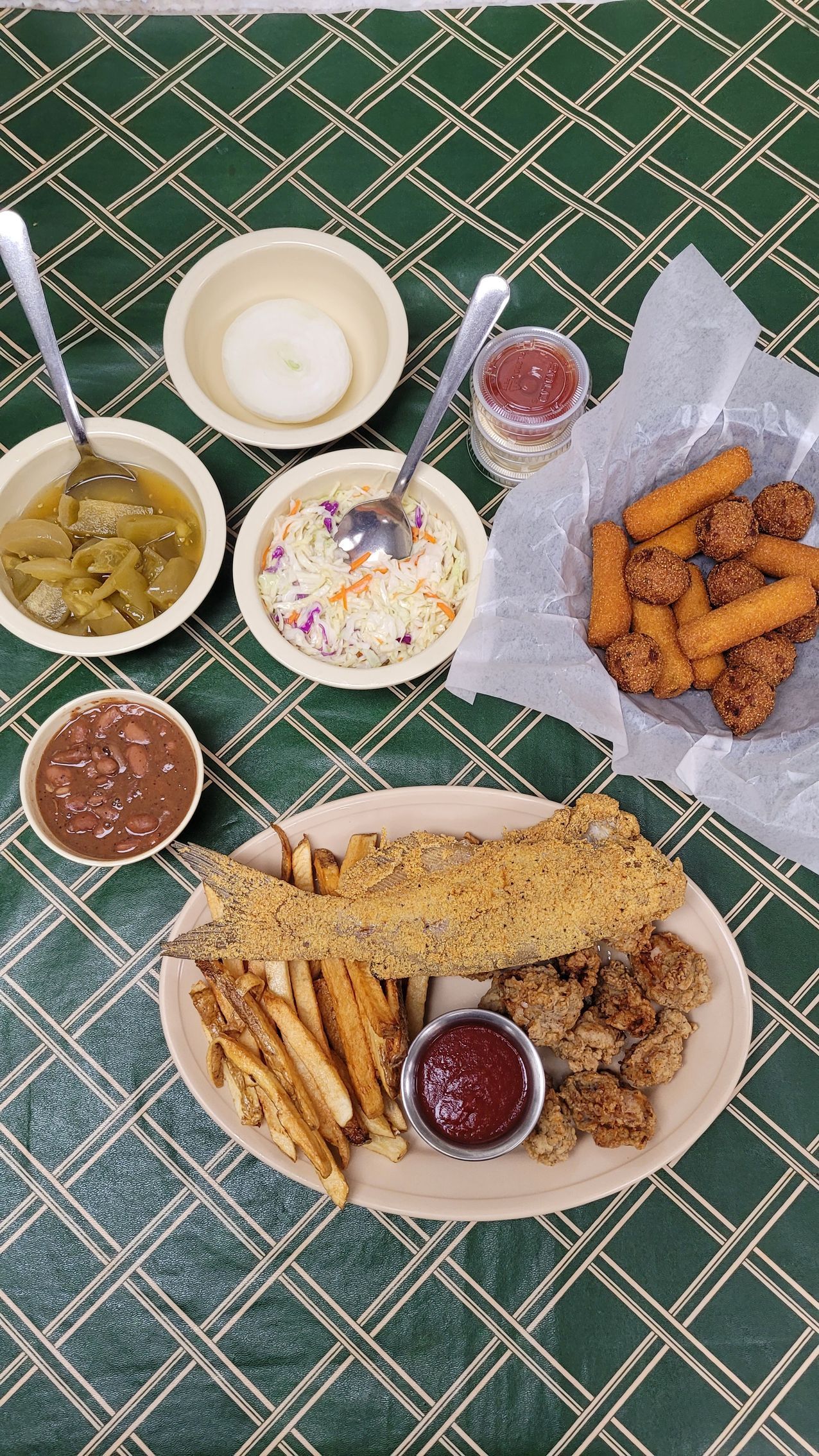A plate of fried food with fries and dipping sauces, alongside side dishes and a basket of fried appetizers on a green checkered tablecloth.