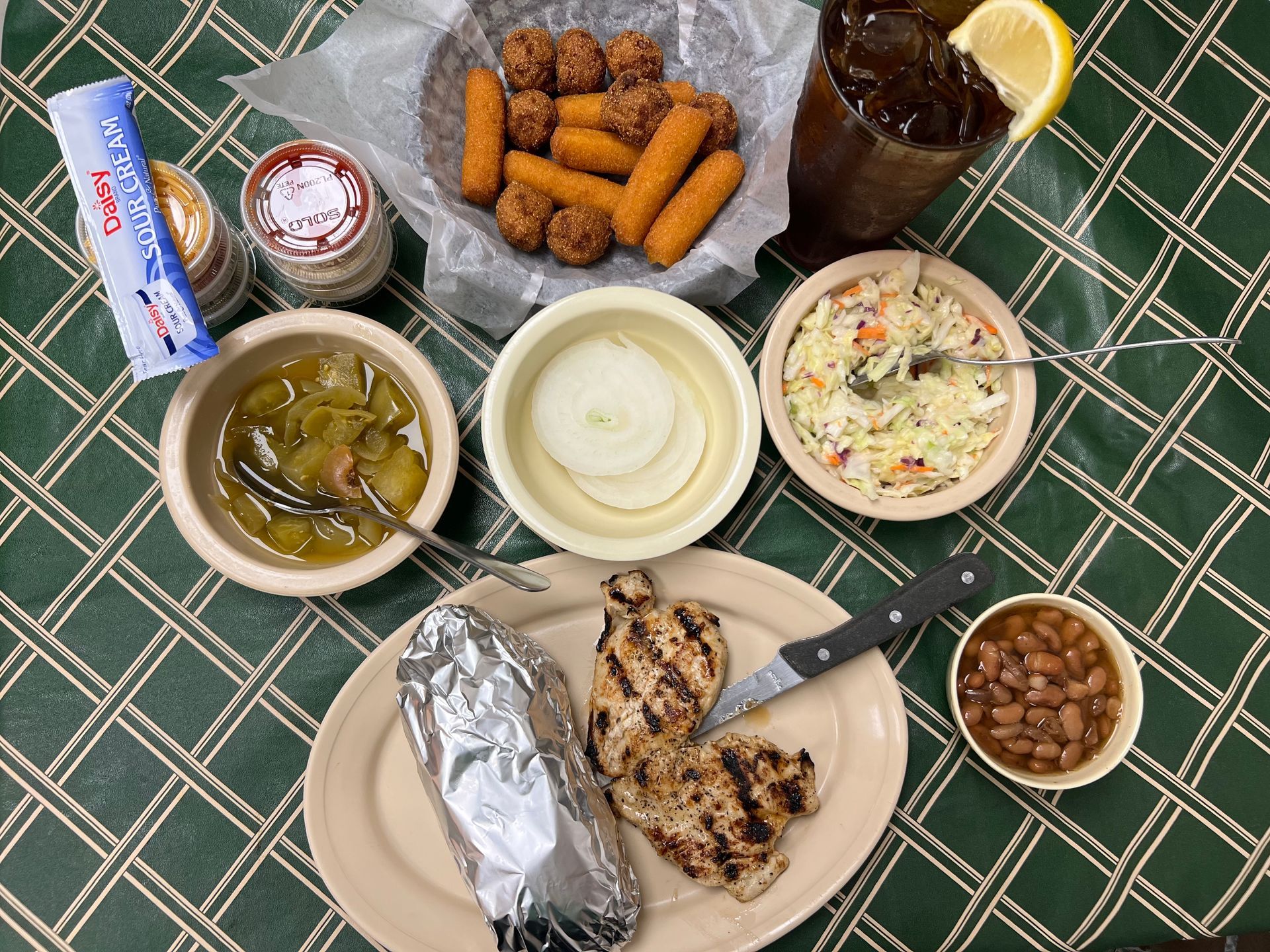 Meal of grilled chicken, baked potato, sides, corn dogs, and iced tea on a green tablecloth.