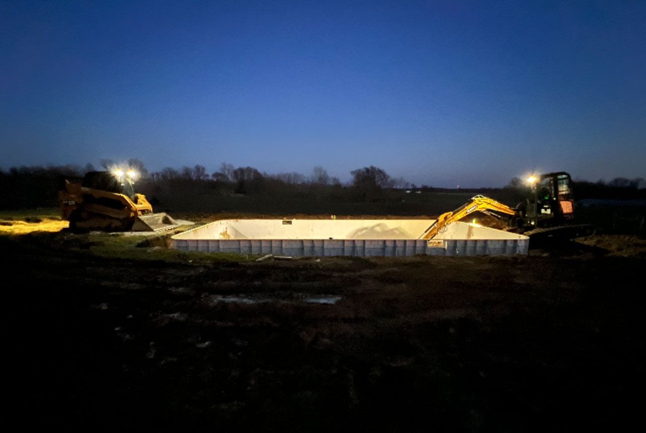 Two excavators illuminate a pool excavation at dusk.