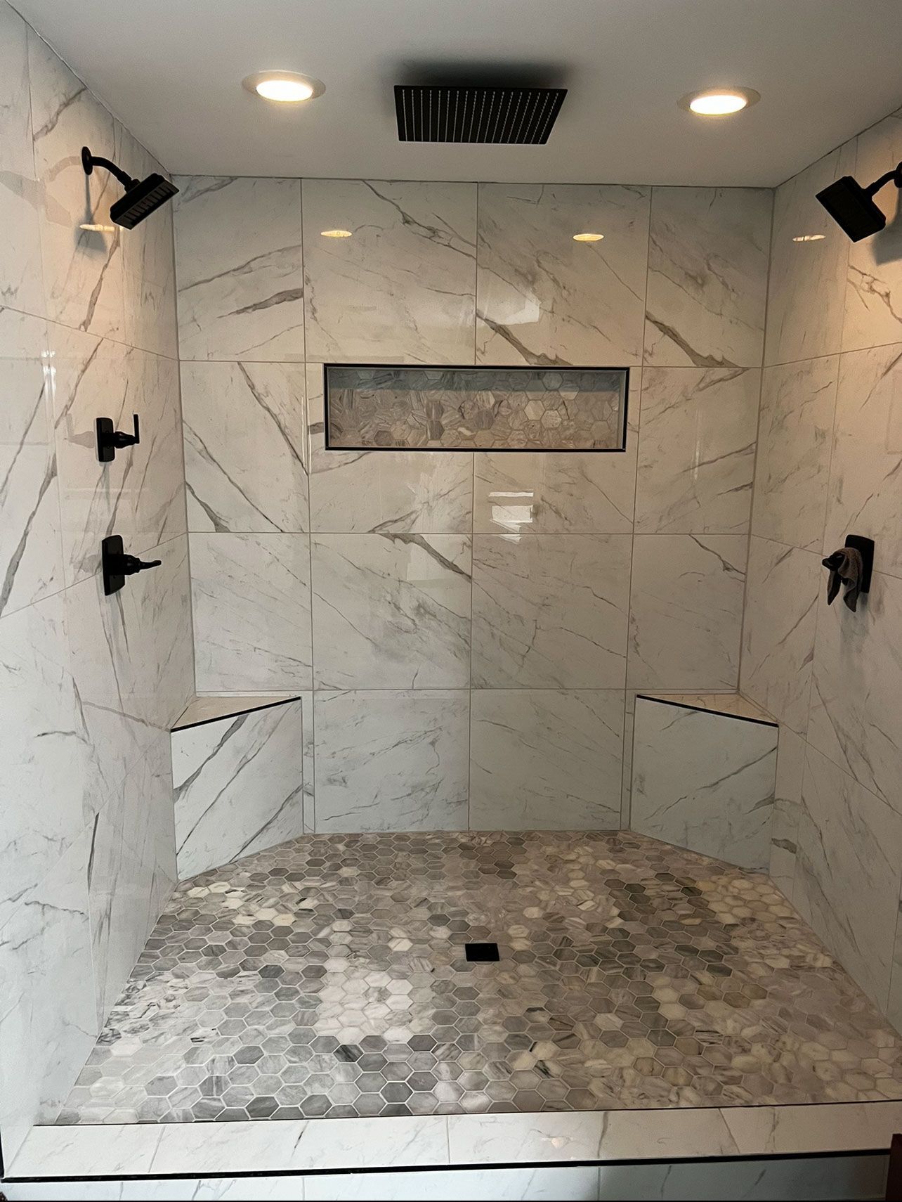Tiled shower with marble-look walls, pebble floor, black fixtures, and recessed shelf.