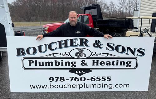 Man standing behind Boucher & Sons Plumbing & Heating sign with phone number and website outdoors.