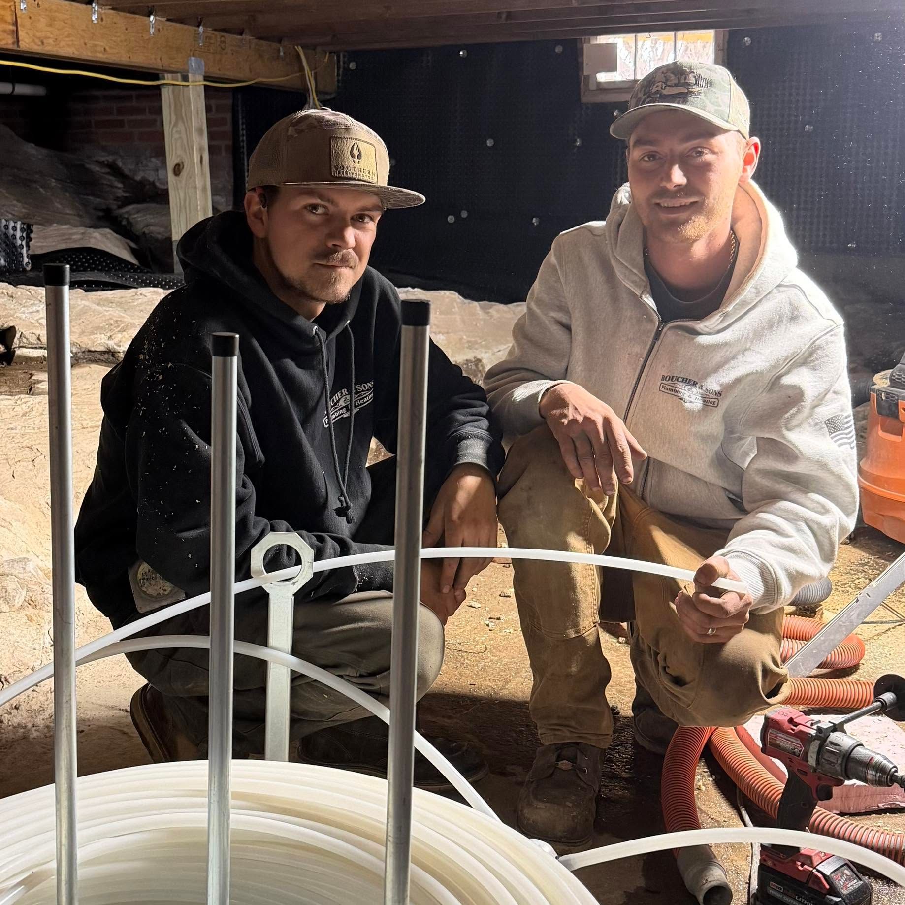 Two men crouch under a table with coiled white tubing in a workshop or garage.