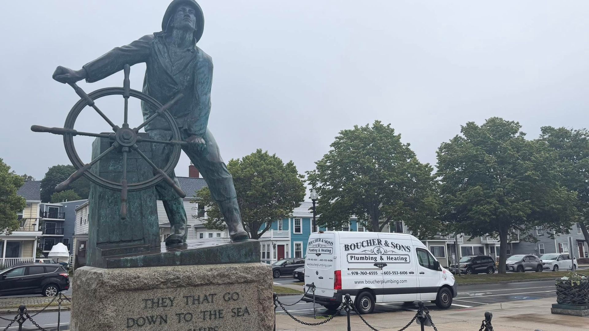 Large statue of a person at a riverside plaza beside a white van, under an overcast sky