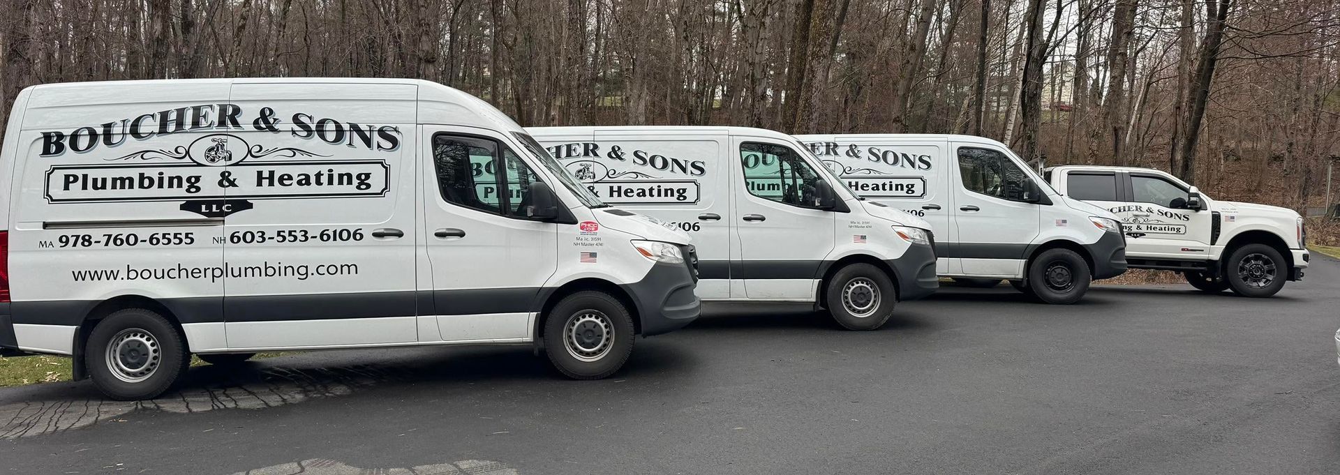 Four white plumbing service vans parked in a row along a roadside by bare trees