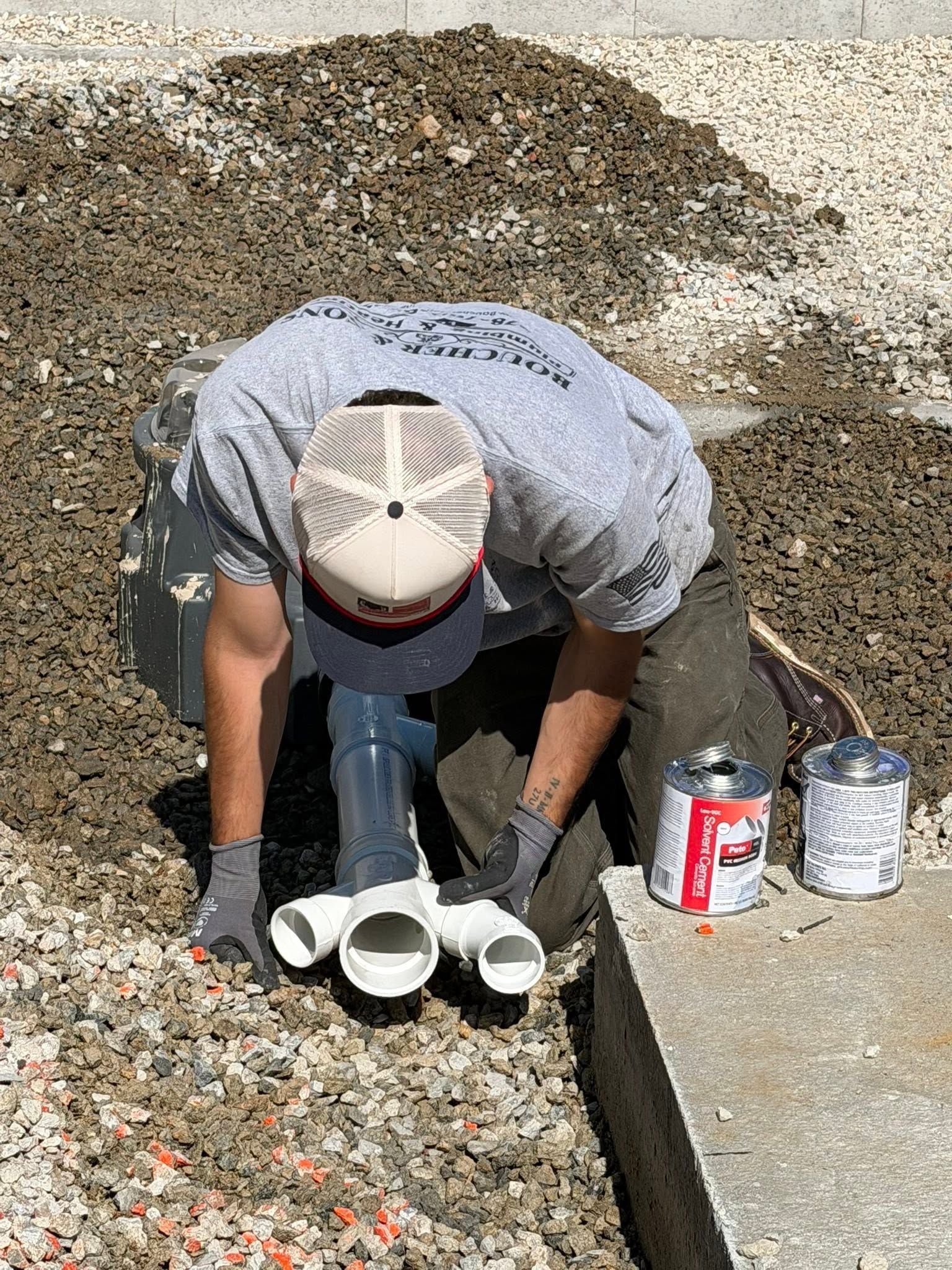 Worker kneeling beside a concrete curb, handling white pipes/tools on gravel near paint cans.