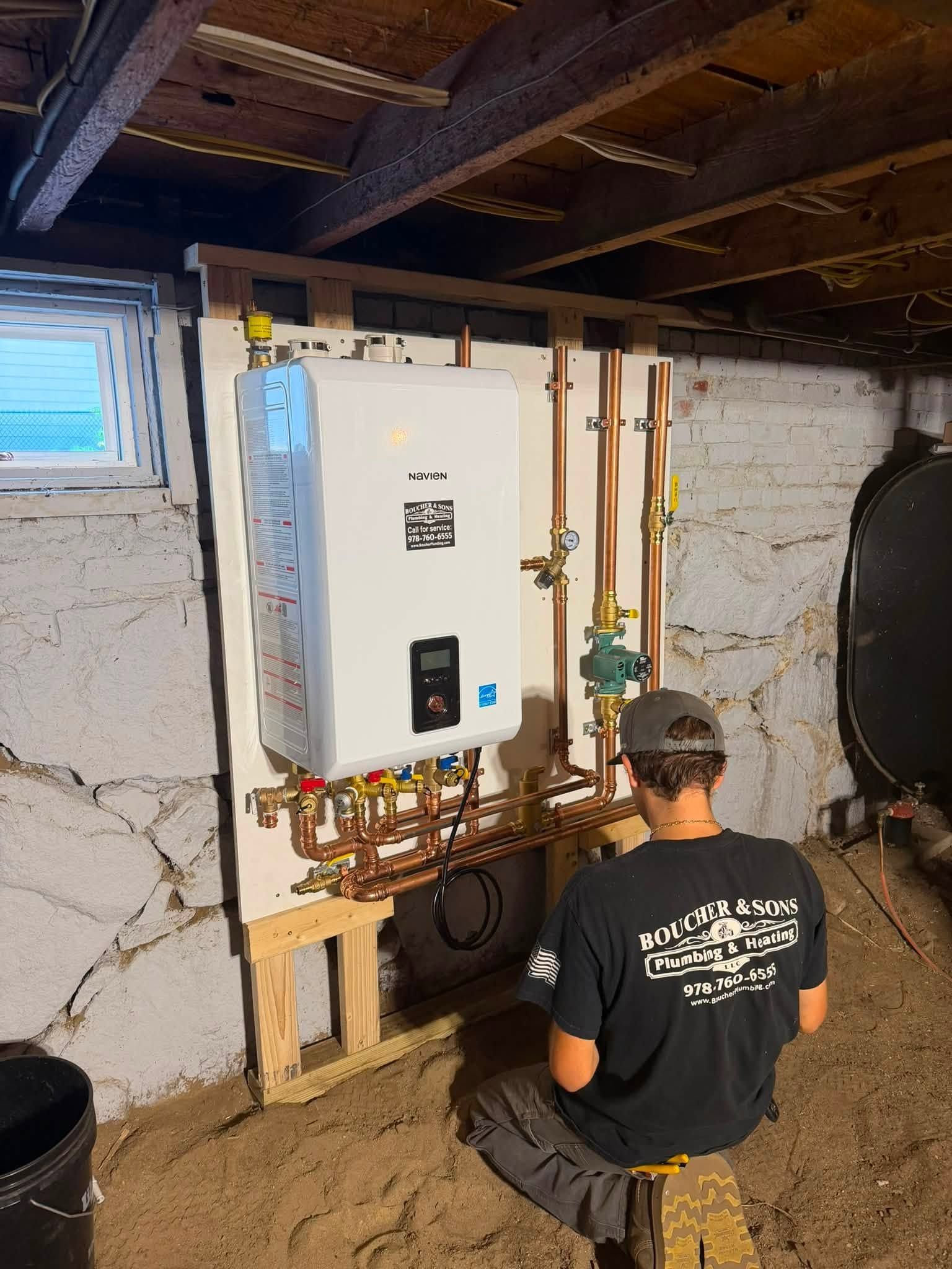 Technician kneeling beside a white wall-mounted boiler and copper pipes in a basement utility area