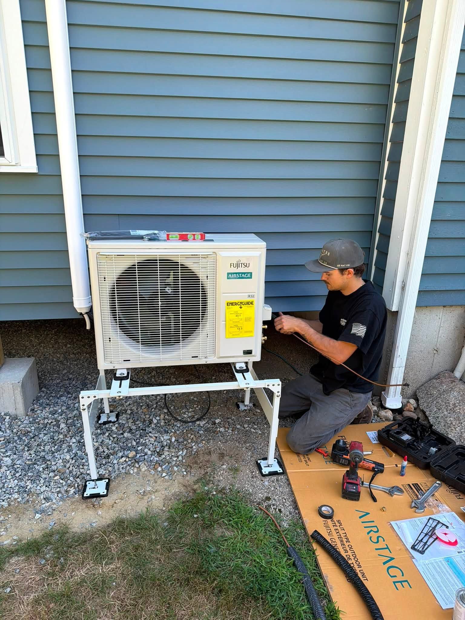 Technician kneels beside an outdoor air conditioner unit, using tools to make repairs by a house siding wall.