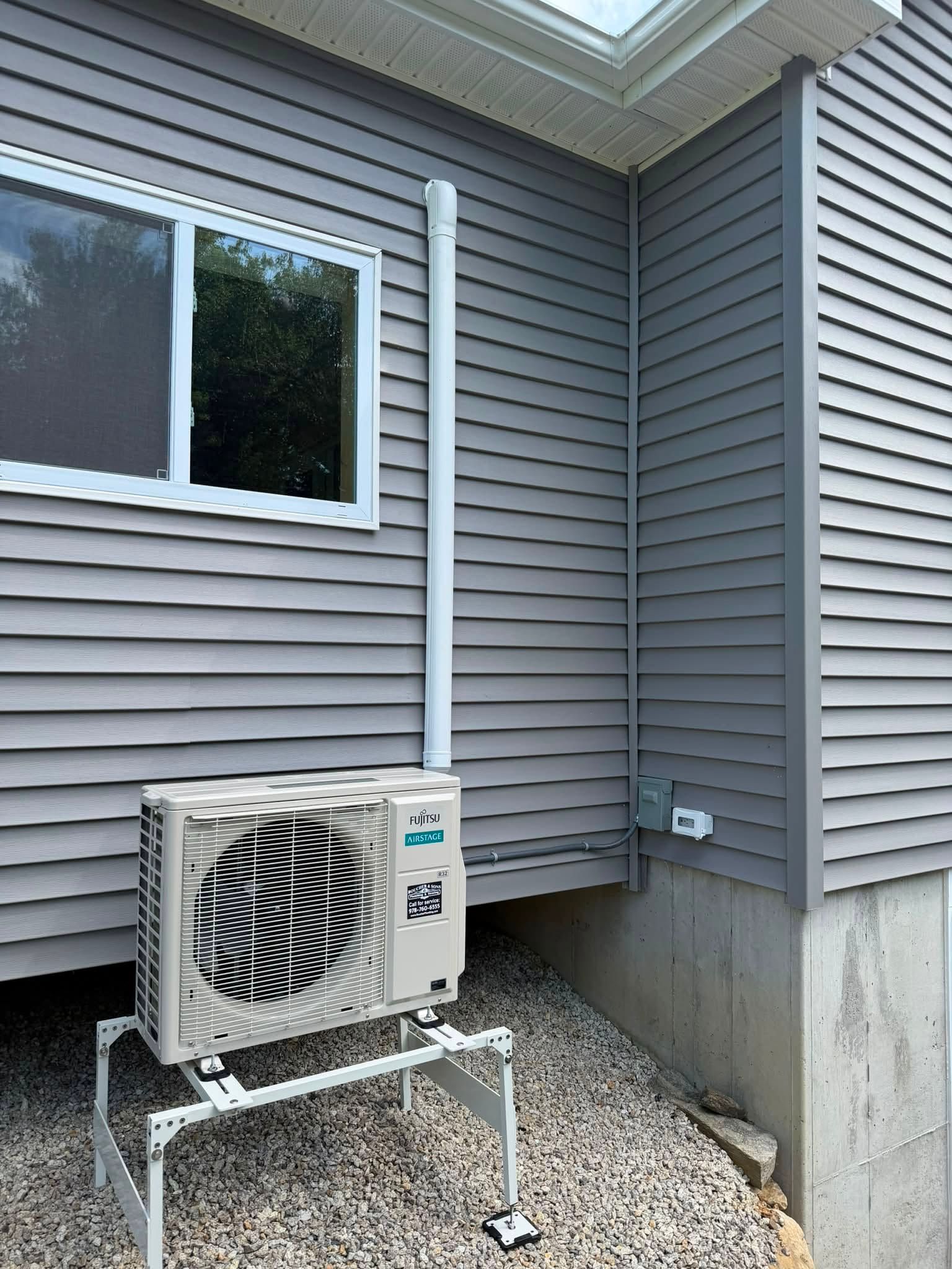 Outdoor air conditioner unit beside a gray vinyl-sided house and window on a gravel bed