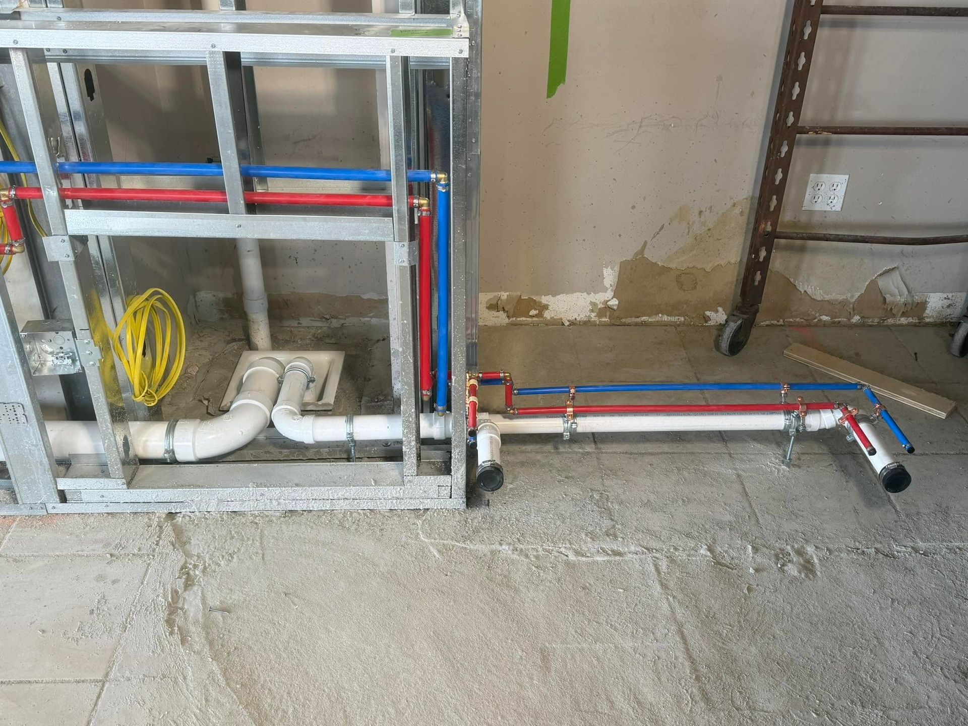 Exposed red and blue plumbing pipes and fittings along a concrete floor and wall inside a construction area
