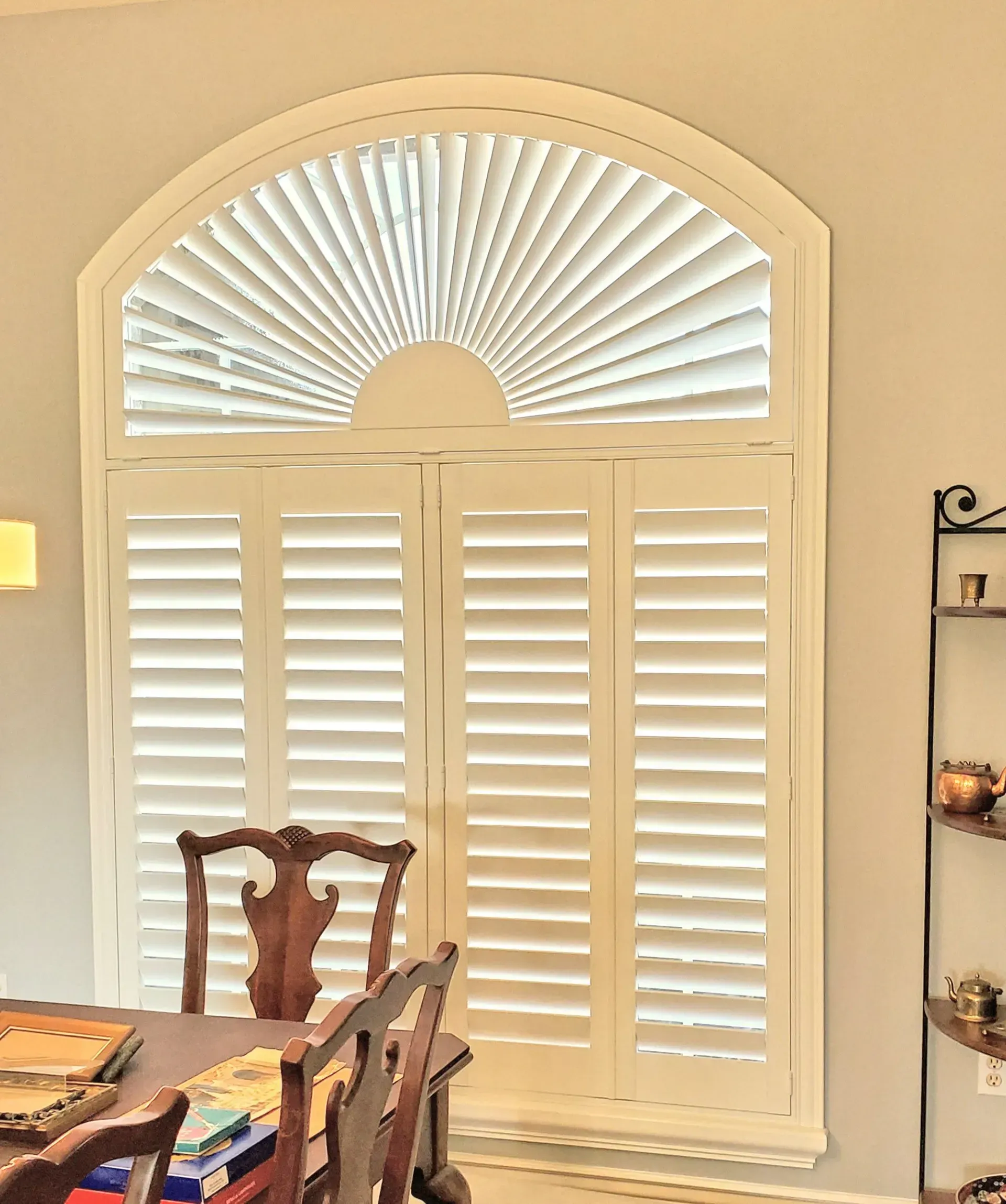 White shutters on a rounded window. Wooden dining table with chairs in front of the window.
