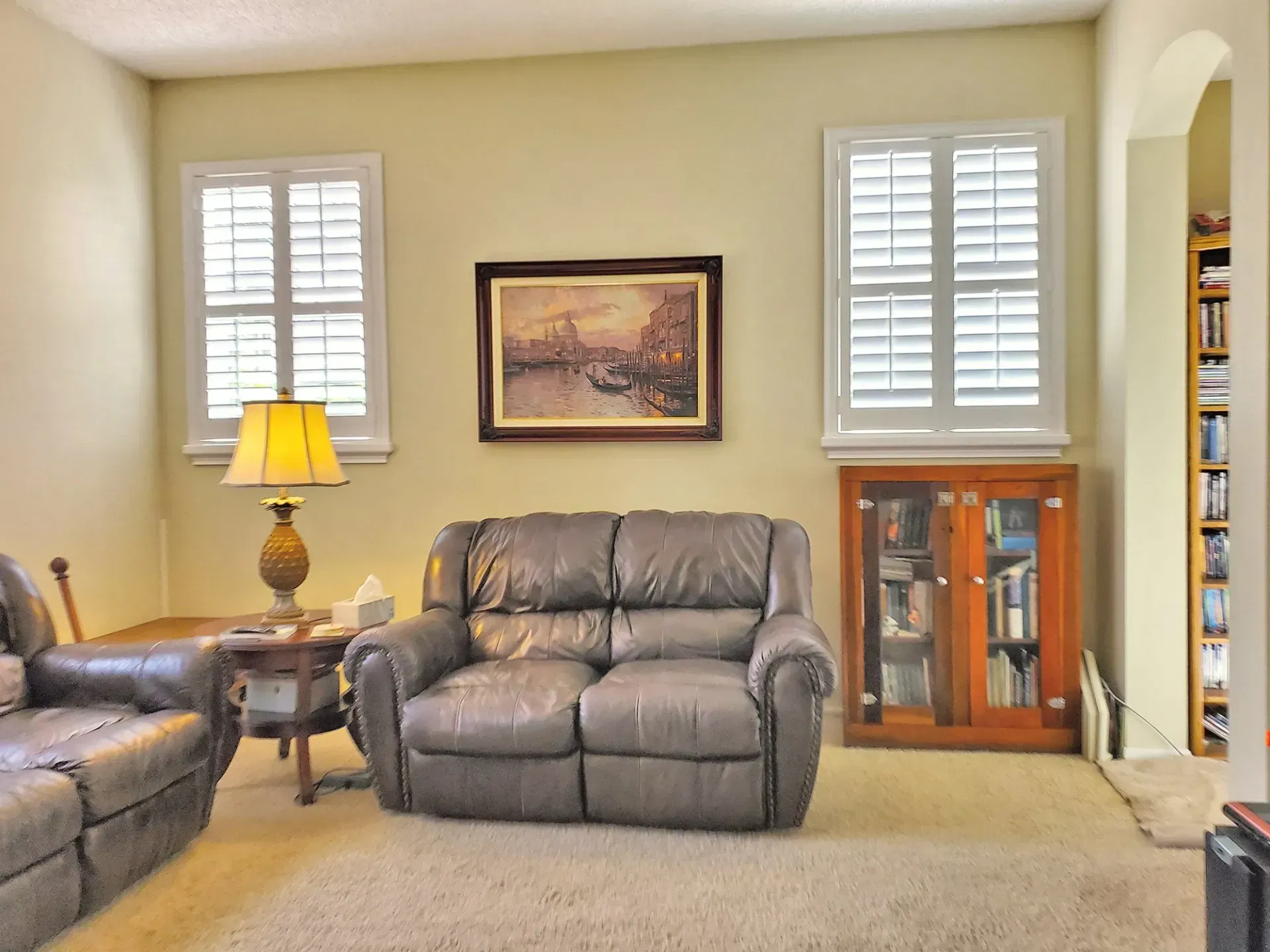 Living room with beige walls, two windows with white shutters, leather furniture, and a painting.