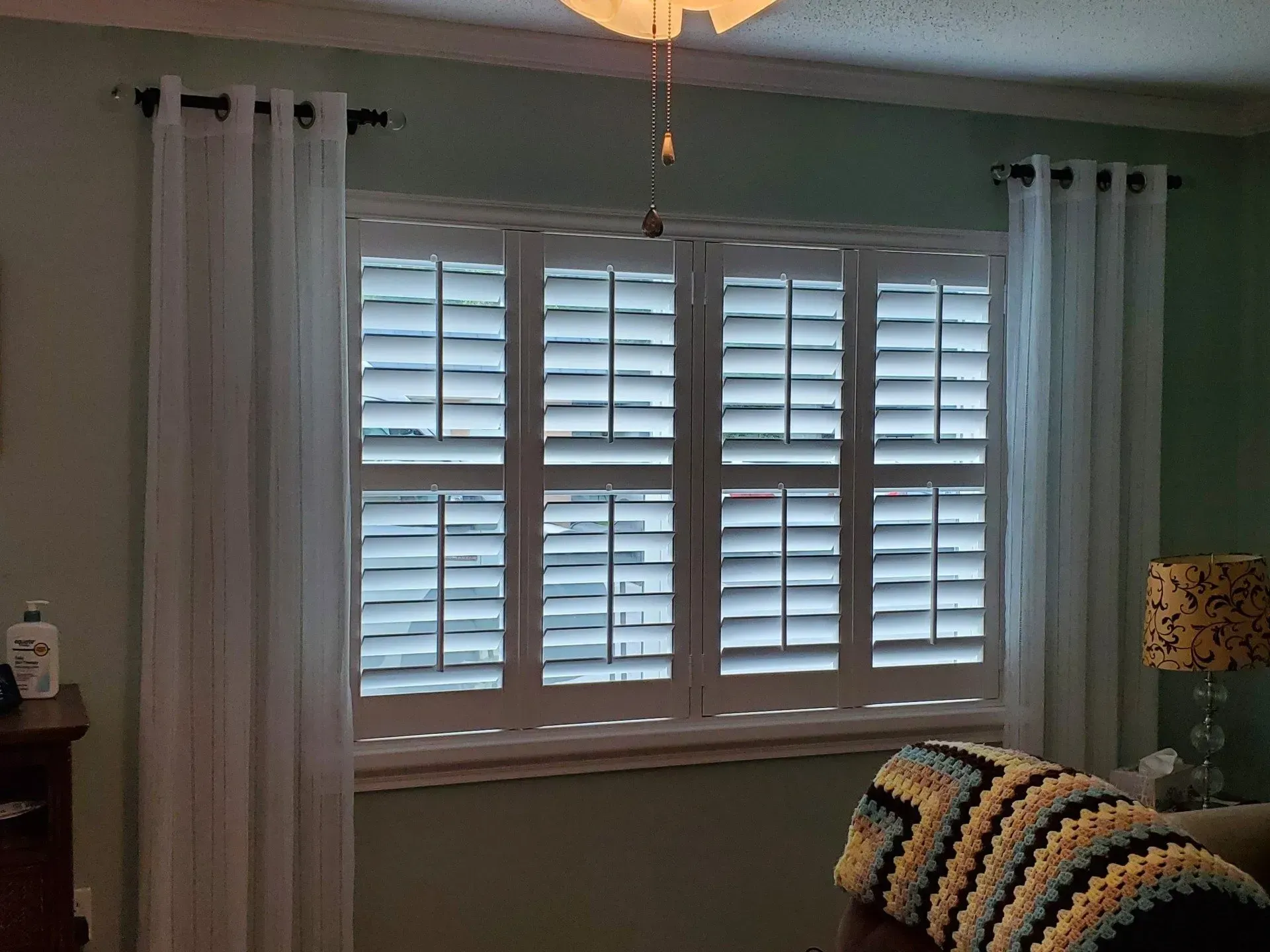 Window with white shutters and sheer curtains, in a room with pale green walls.