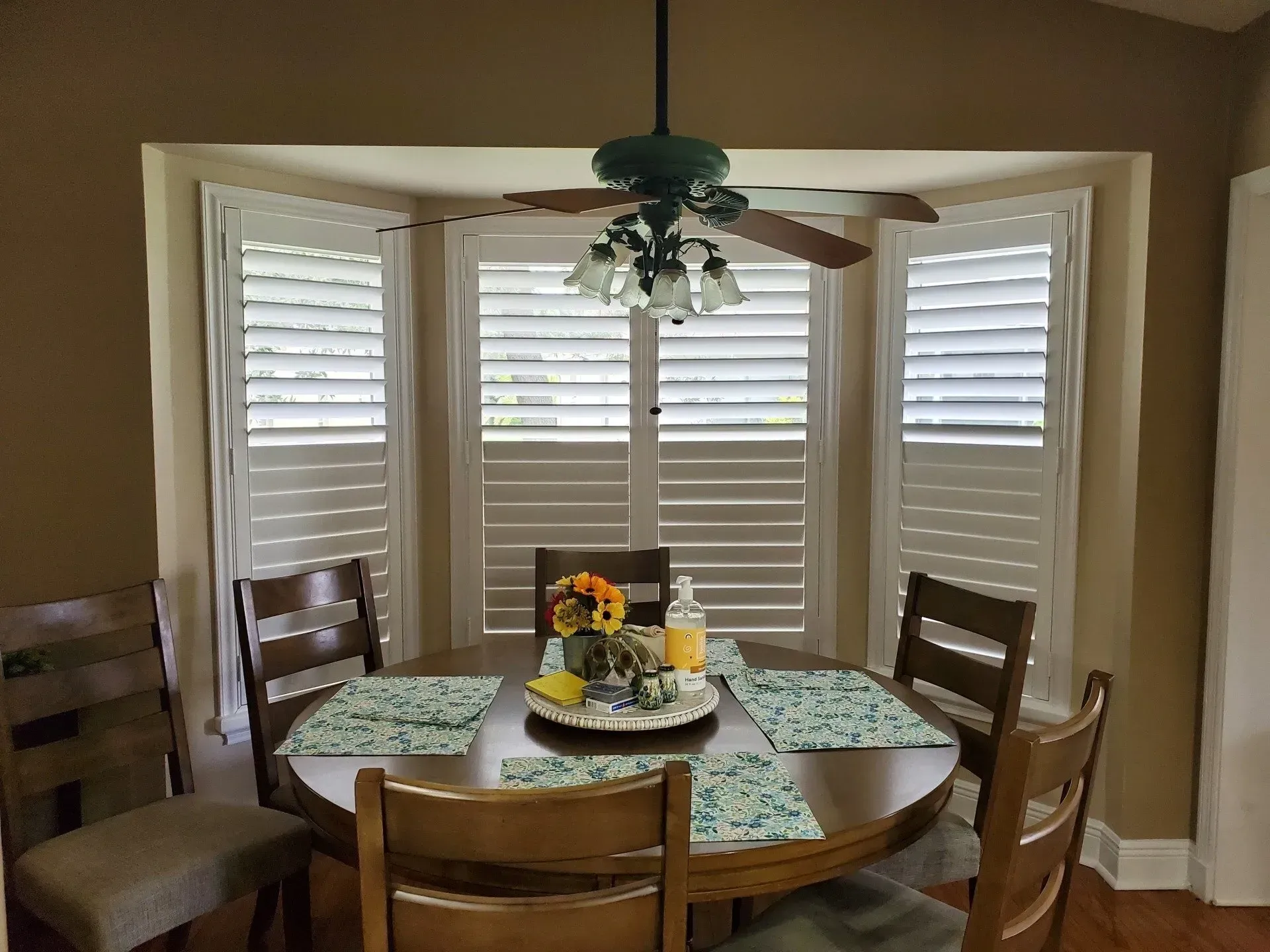 Dining table set for four in front of a bay window with white shutters. A ceiling fan hangs above.