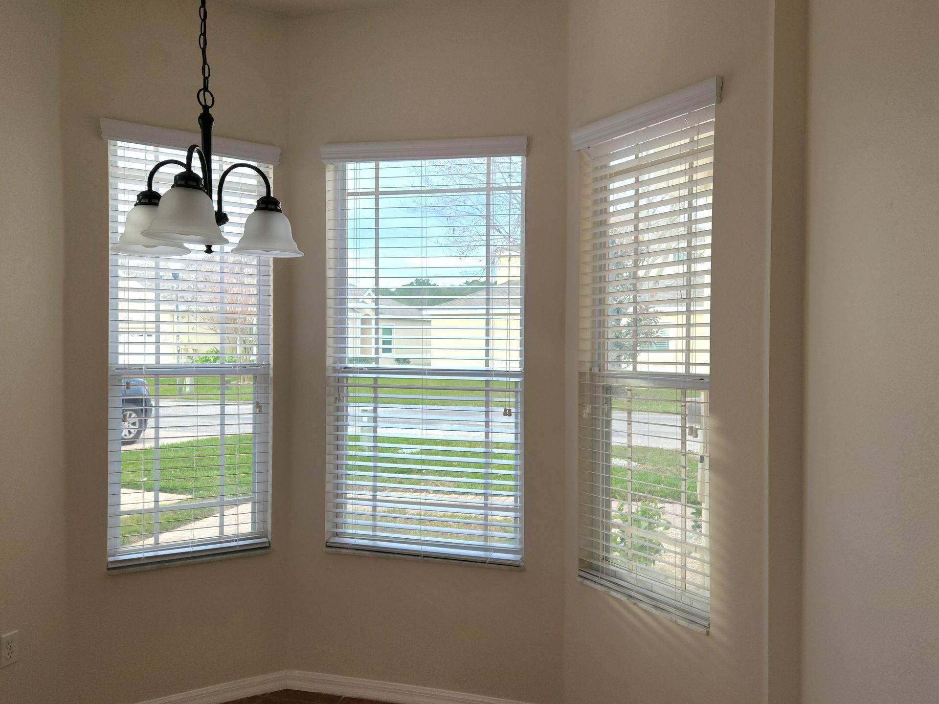 Three windows with blinds in a room; a chandelier hangs above them. Beige walls.