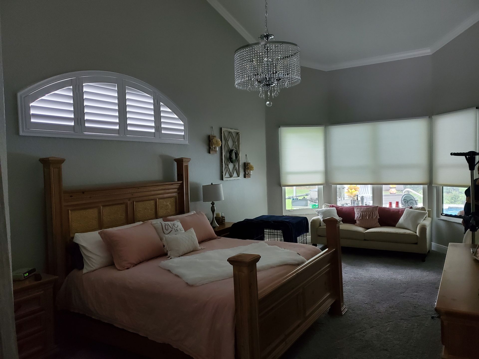 Bedroom with light gray walls, wooden bed and furniture, arched window shutters and a chandelier.