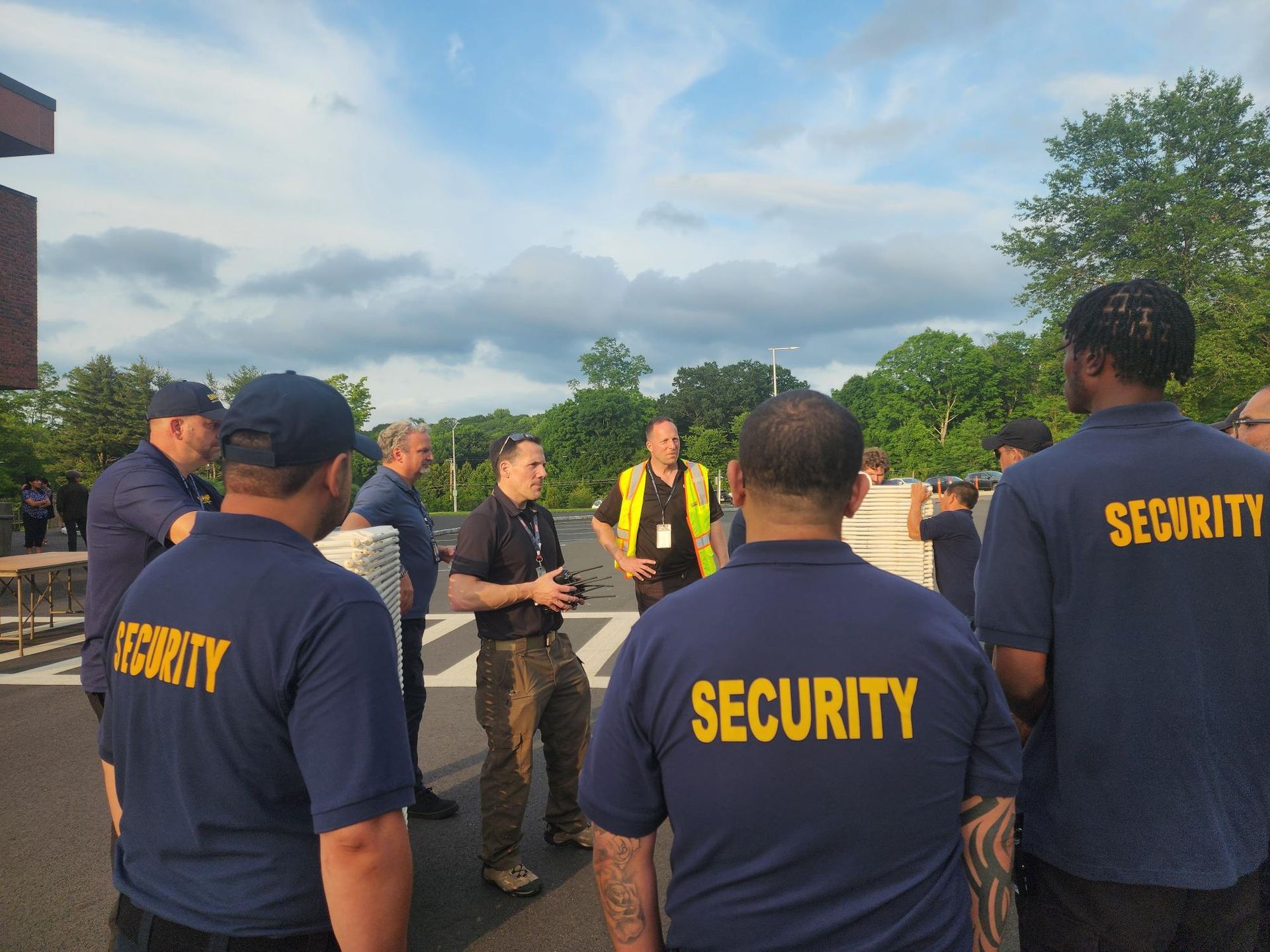 Security staff stand in a road listening to a speaker at sunset, with trees and a cloudy sky behind them.