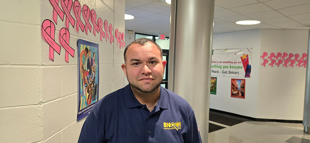 Man in a blue shirt in a school hallway with pink ribbons and posters on the wall.