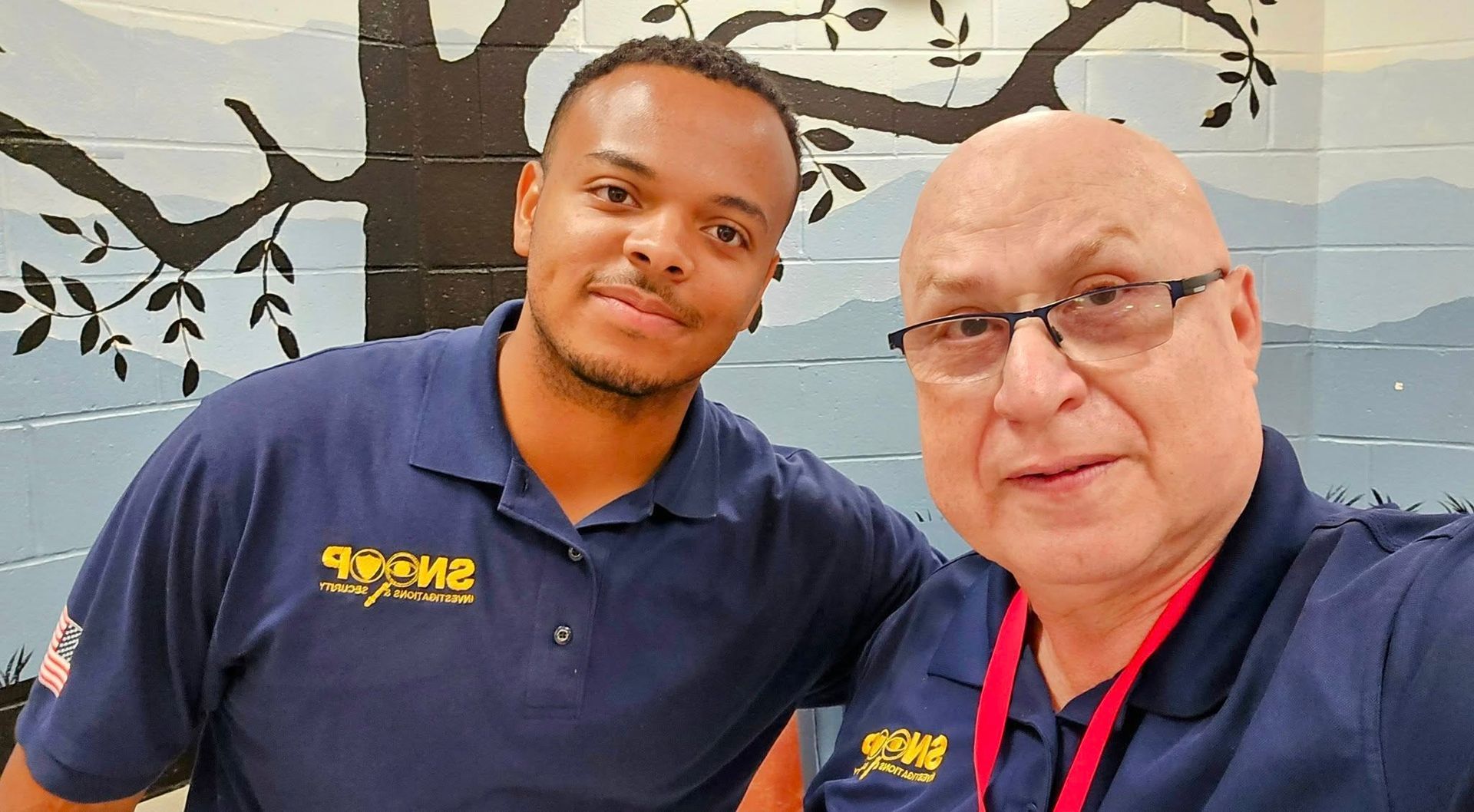 Two men posing indoors, wearing navy polo shirts, with a wall mural in the background.