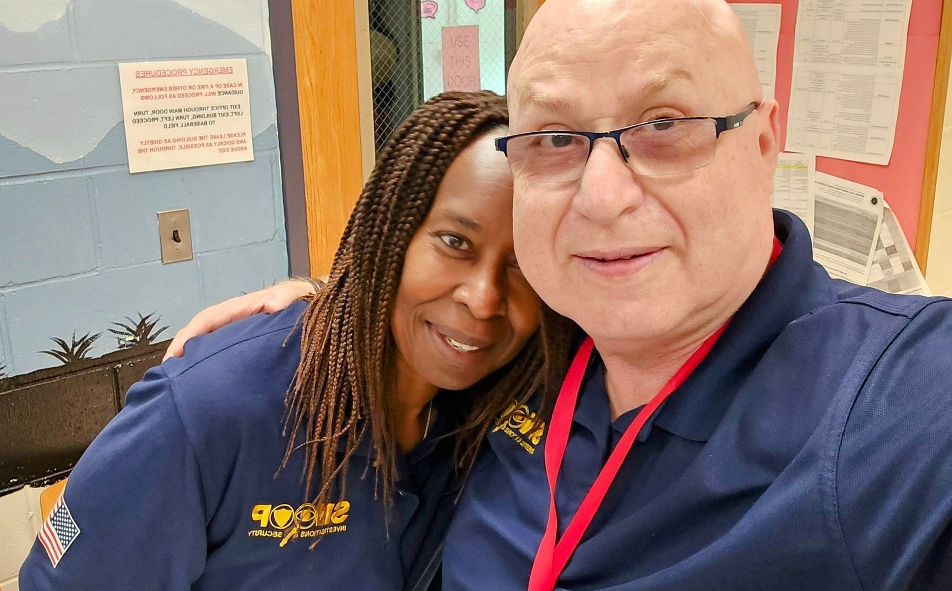 Two people smiling close together indoors, wearing navy shirts and a red lanyard