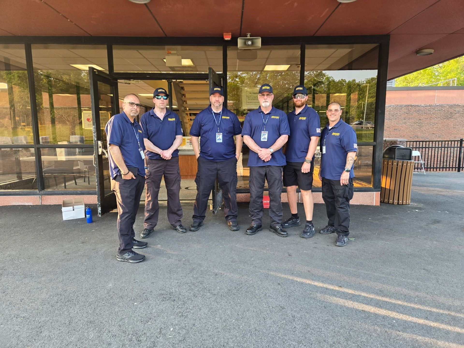 Six volunteers in matching blue shirts stand outside a building entrance, posing for a group photo.