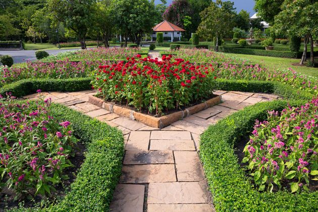 A sunny, formal garden with a stone path leading to a central raised flower bed of red celosia surrounded by purple flowers.