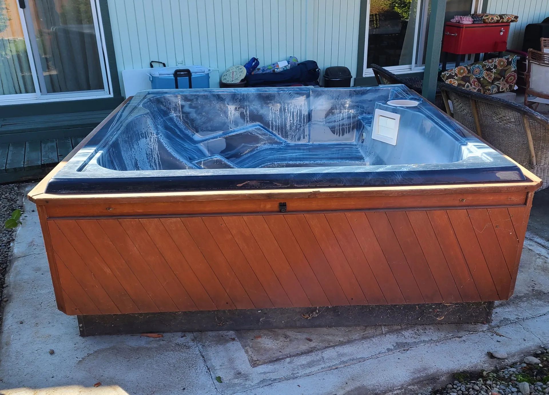 Wooden outdoor hot tub with blue cover, sitting on a concrete patio beside a house